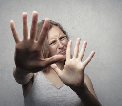 Portrait of a fearful woman in a gray tank top with hands pushed forward against a gray background.